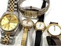 Photo showing all five wrist watches arranged on a white surface, displaying various styles and bands, from metal linked bracelets to leather straps.