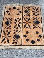 Full view of folded tapa bark cloth with brown and black geometric and botanical painted motifs on tan background, showing two main vertical panels.