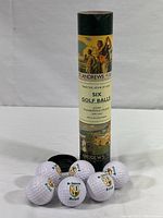 Set of six golf balls with printed St. Andrews Scotland crests arranged around the bottom of the open cylindrical tube container with golf artwork label, placed upright on a white surface.