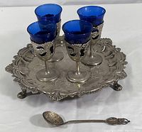 Four blue cobalt cordial glasses with ornate silverplate holders and stems placed on an ornate silverplate tray, with a small stir spoon in front.