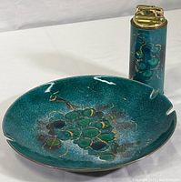 Photo of circular enameled brass ashtray with green grape cluster design beside matching lighter