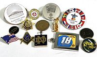 Top view of various vintage pins and tokens arranged on a white surface, showing logos and emblems like Burning Man, Coca-Cola Olympics, and Callaway Golf.