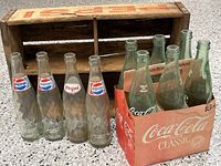 Photo showing six Coca Cola Classic 16 oz glass bottles in faded red cardboard carrier alongside four 12 oz Pepsi bottles and a wooden Pepsi bottle carrier with loose slats.