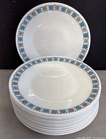 Stack of 12 white ceramic plates with geometric blue and black pattern on rim.