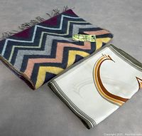 Pair of scarves side by side on a flat surface, showing colorful zigzag scarf with fringe and white scarf with gold pattern folded.