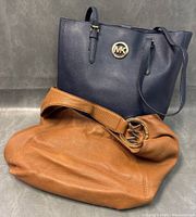 Two Michael Kors handbags shown together on grey background, one navy blue tote and one tan hobo bag.