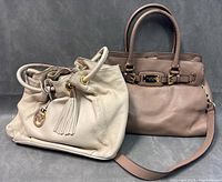 Two Michael Kors handbags together; beige drawstring style and taupe satchel style.