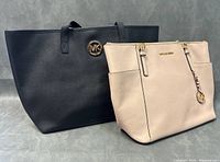 Pair of Michael Kors handbags: a black tote bag with gold MK emblem and a beige crossbody bag with gold logo charm and nameplate.