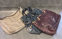 Three Michael Kors designer bags: beige shoulder bag, black tote with chain detail, and burgundy textured handbag arranged on a grey surface.