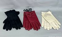 Three pairs of gloves displayed side by side: black suede gloves, red leather gloves with bow, cream leather gloves with decorative stitching