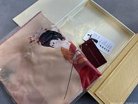 Silk scarf with Geisha design, shown folded inside transparent zip pouch and with attached brand tag.