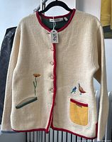 Front view of cream wool jacket with red trim, two front pockets, embroidered flower on left pocket and embroidered birds on right pocket.
