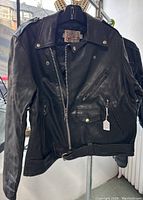 Front view of black leather jacket hanging, showing zippered pockets, belted waist, and snap collar