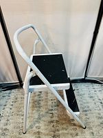 Front angled view of the 2-step foldable ladder showing white metal frame, black steps and curved safety handle.