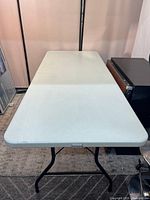 Front angled view of white rectangular plastic folding table with black metal legs and adjustable height.