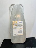 Photo of folded countertop ironing board with iron rest on black surface against white wall, label showing product info and brand 'today'