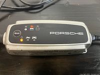 Close-up of Porsche branded Charge-o-Mat Pro battery maintainer charger with control lights and mode button visible.