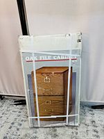 Photo shows the original packaging of the oak file cabinet with image of the cabinet printed on the box. The packaging is strapped and intact with some minor cardboard damage visible.