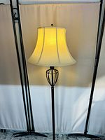 Front view of the standing lamp with illuminated cream fabric shade showing ornamental metal frame beneath shade.