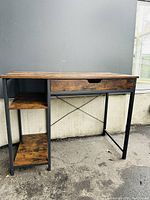 Front view of desk showing wooden top, drawer with cut-out handle, two open shelves on side, and black metal frame Structure.