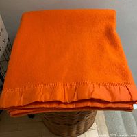 Folded tangerine wool blanket with satin binding and blanket stitch edges, sitting on a basket.
