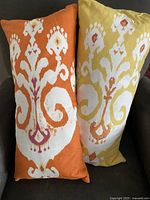 Front view of two rectangular cotton pillow covers with feather pillows inside. One is tangerine orange with a large white ornamental pattern accented with maroon and yellow. The other is butter cream yellow with a matching pattern in white and accents of maroon and yellow.