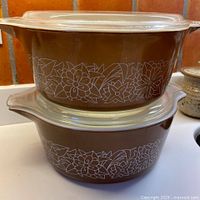 Two brown Pyrex casserole dishes stacked on top of each other showing white floral and leaf pattern on a brown background.