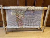 Full front photo of the needlepoint piece on the wooden adjustable rotating frame showing the girl with curlers and teddy bear design and chevron pattern top edge.