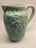 Full side view of pale minty green/blue ceramic pitcher displaying raised foliage and animal motifs.