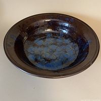 Side angled view of a large studio pottery bowl showing dark chocolate brown glaze with mottled blue/grey detailing inside and blue glaze drip on side.