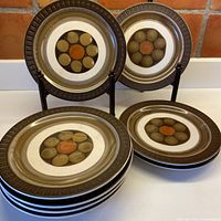 Group photo showing 9 stoneware plates in the Mirage pattern with chocolate brown rims, olive green, cream, and tangerine center decoration.