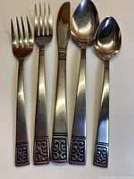 Photo showing sample place setting of stainless steel flatware with detailed motif on handle ends including knife, dinner fork, dessert fork, soup spoon, teaspoon.
