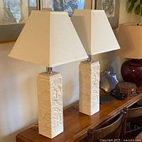 Two vintage square ceramic lamps with detailed fossil designs and creamy beige lamp shades, shown on a wooden surface in a room with framed art on the wall.