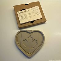 Photo showing heart-shaped ceramic cookie mould with maple leaf relief alongside original packaging and instruction booklet.