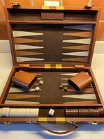 Opened backgammon set showing full game board with pointed pattern, 15 brown checkers and 15 cream checkers lined up, two brown dice cups, two sets of dice, and doubling cube in center.