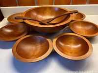 Full set of one large maple bowl, six smaller bowls, salad spoon and fork arranged on surface.