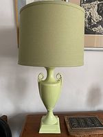 Soft green ceramic lamp with urn-shaped body, curved foliage handles, and square base shown with new lampshade on a wooden surface against a wall