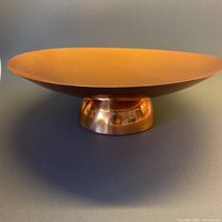 Full side view of the round shallow copper bowl on stem base against dark neutral background