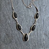 Full view of the sterling silver necklace showing five pear-shaped onyx stones bezel set in silver, laid flat on dark slate background
