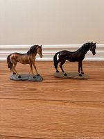 Two horse figurines side by side on wood floor against white baseboard and wall.