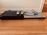 Two RCA DVD players side by side on wooden floor with two remote controls on top of one player.