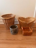 Photo of two wicker baskets, one small basket with handle, and one gray clay plant pot on wooden floor against white wall.