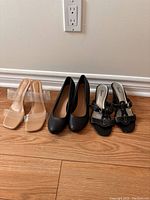Frontal view of three pairs of women's heeled shoes including clear heel sandals, black wedge heels, and black buckle sandals.