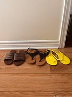 Three pairs of women's sandals lined up on wooden floor showing yellow flip flops, black T-strap sandals and brown woven slide sandals.