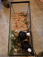 Top view of the aquarium tank showing wood shavings substrate and two black heat lamps with white sockets, as well as artificial greenery inside the tank.