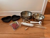Photo showing the 2 heart-shaped cake trays in black metal, 3 round openable metal cake trays, a white Braun handheld mixer with beaters, and red plastic cookie cutters.