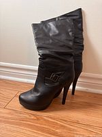 Side profile view of single black heel boot with slouched shaft, decorative buckle strap and high stiletto heel on wood floor.