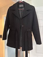 Front view of the black wool dress coat hanging on a pink hanger, showing button closure, collar, and flap pockets.