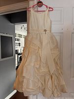 Front view of ivory flower girl dress on hanger, showing layered gathered skirt with decorative bow and brooch detail.