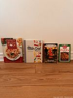 Front view of four cookbooks arranged side by side on a wooden floor against a white wall.